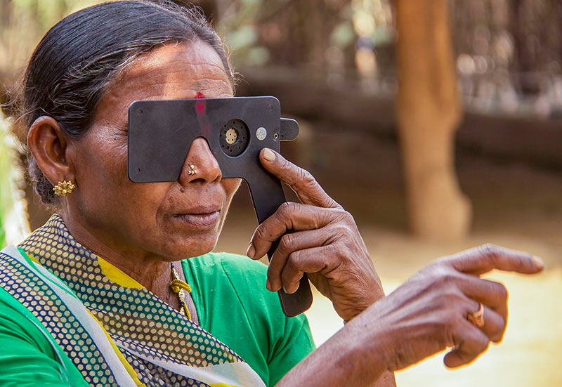 A lady holds a pin hole device with her left hand to her left eye and points forward with her right hand