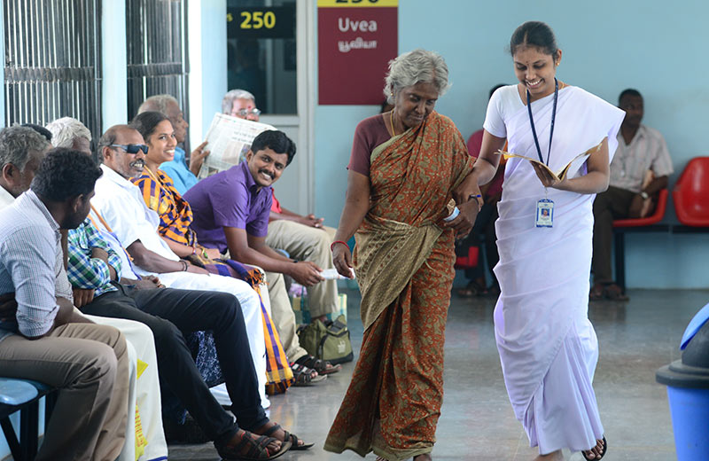 A female eye hospital staff member takes the arm of an elderly female patient to steady her as they walk together along a hospital corridor, past several people sitting down on chairs waiting for appointments