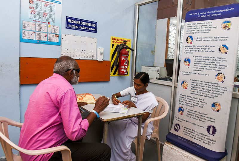A female pharmacist is sitting across the table from a male patient. She is writing.