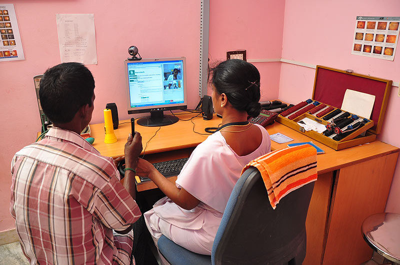 Two eye health workers, one male and one female, sitting at a desk in front of a computer in a refraction room