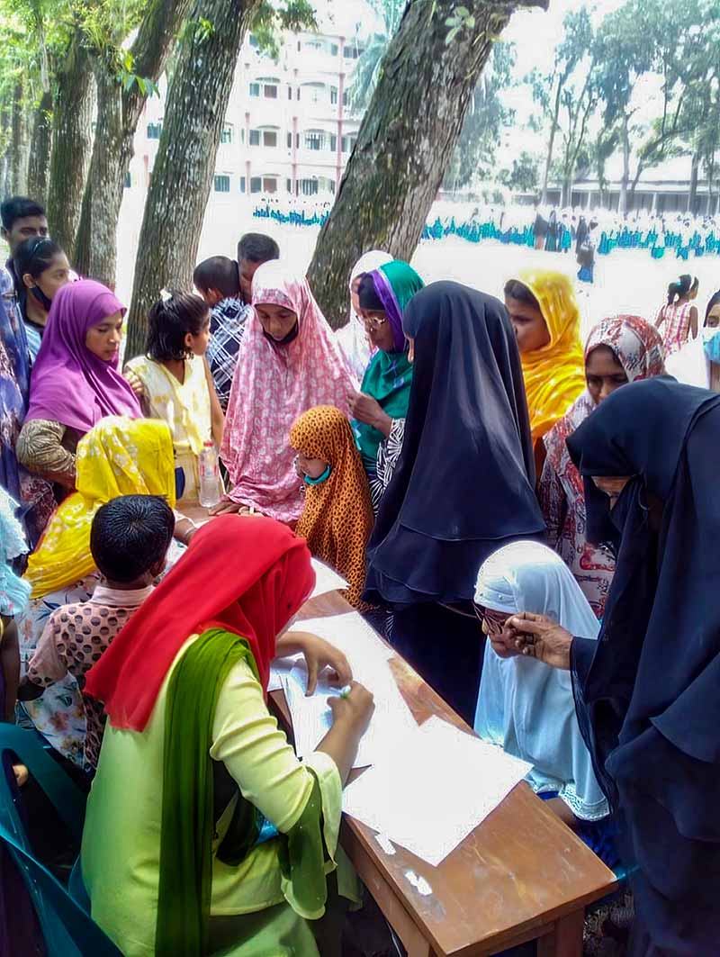 Several women and girls stand in front of a registration desk under some trees.