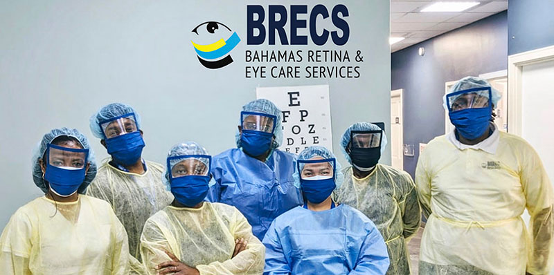 Seven health care workers wearing PPE (gowns, hair caps and visors) stand together.