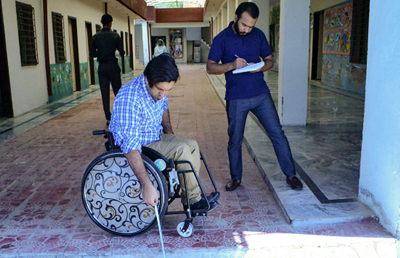 A man writes down notes on a pad of paper. He is standing next to a man sitting in a wheel chair.