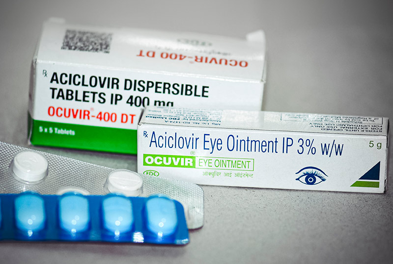 Photographie montrant dans le même cliché de l’aciclovir sous plusieurs formes : une boîte de comprimés d’aciclovir, une boîte contenant de la pommade à l’aciclovir, et deux plaquettes montrant des comprimés d’aciclovir sortis de leur boîte.