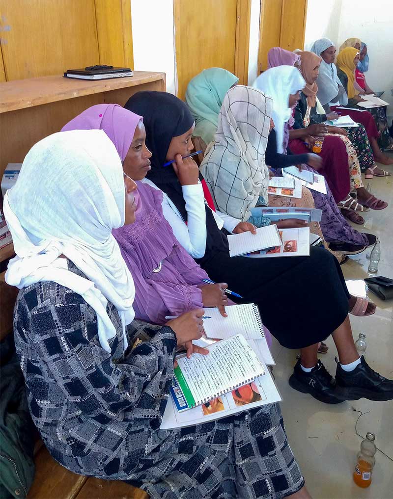 A group of women sitting in a row on a bench. Each woman is holding a notepad, pen and printed literature.