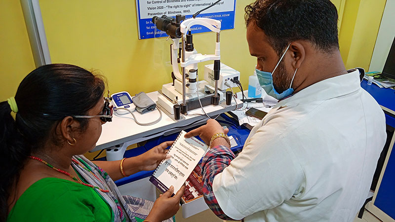 A male optometrist assists a female patient by pointing out rows of text on the near vision chart she is holding