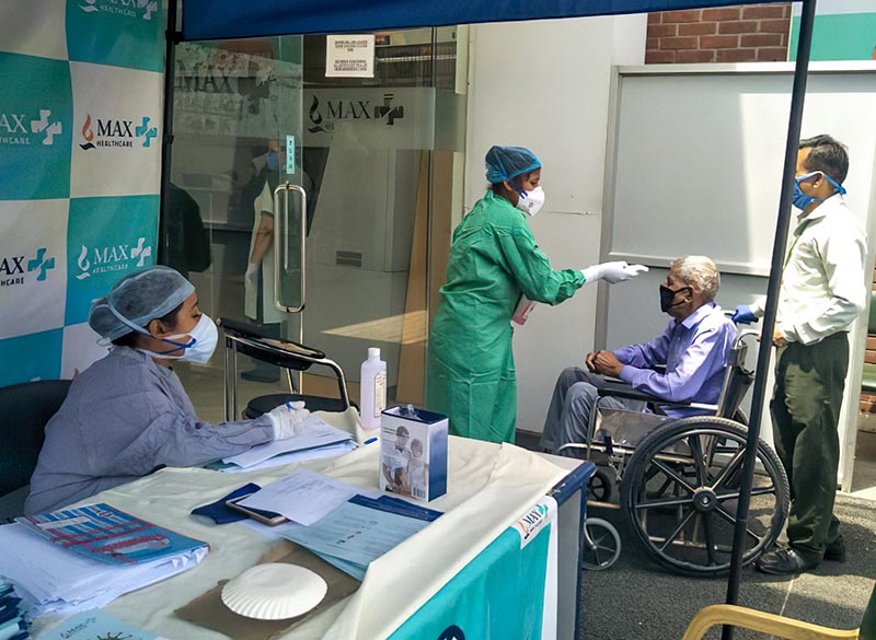 A patient in a wheelchair is being accompanied to the clinic where a female health worker checks his temperature at the entrance