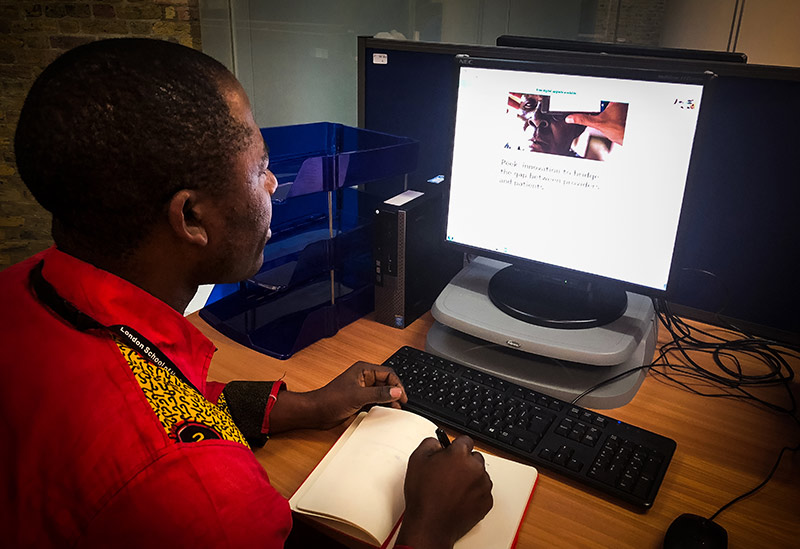 Man sitting at a desk looking at a desktop computer screen, writing notes with a pen and paper in a notebook