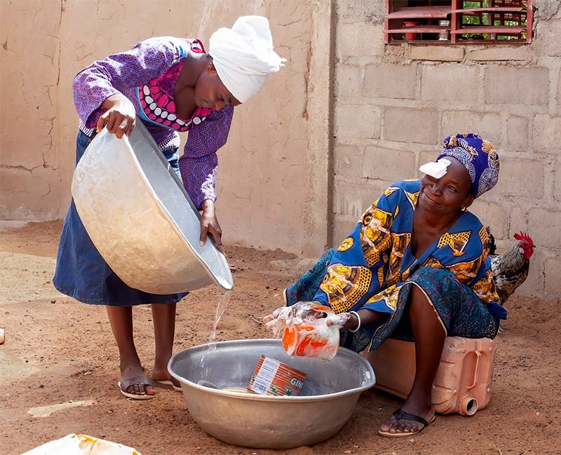 Two ladies in a a yard outside a house. One lady is holding a large metal bowl fro which she is pouring water into another large metal bowl on the ground in front of a lady who has a bandage over her right eye.