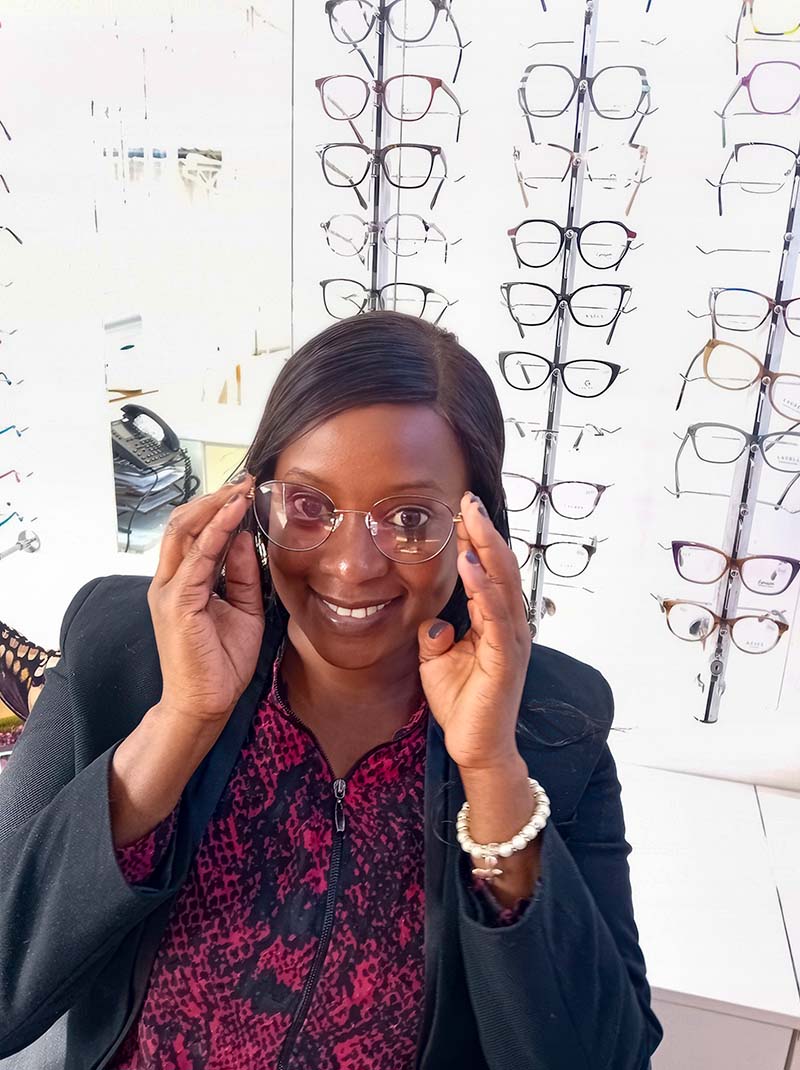 A woman tries on a pair of spectacles. There are glasses mounted on the wall behind her.