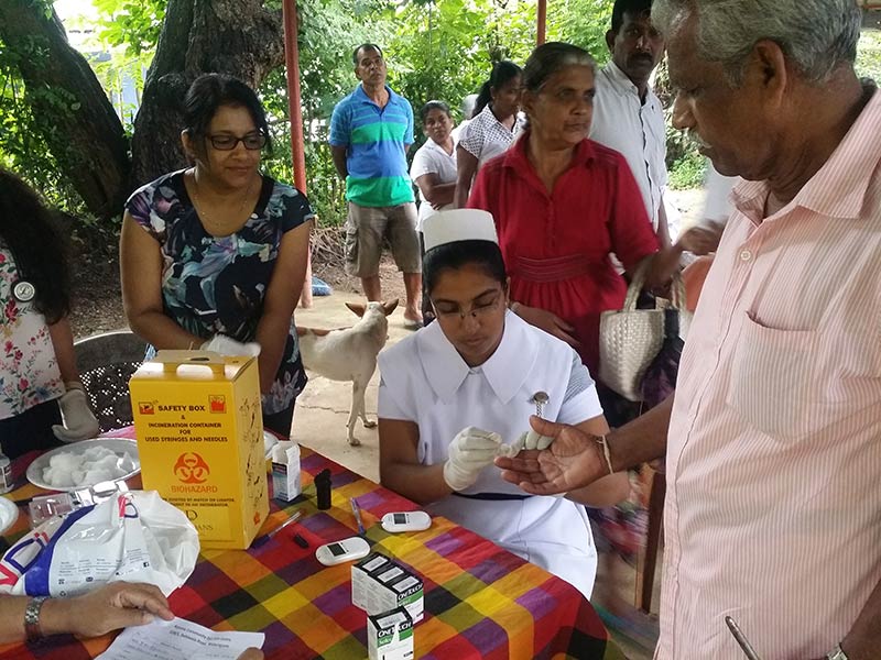 Nurse working from a table outside, taking blood via finger prick, surrounded by people and a dog