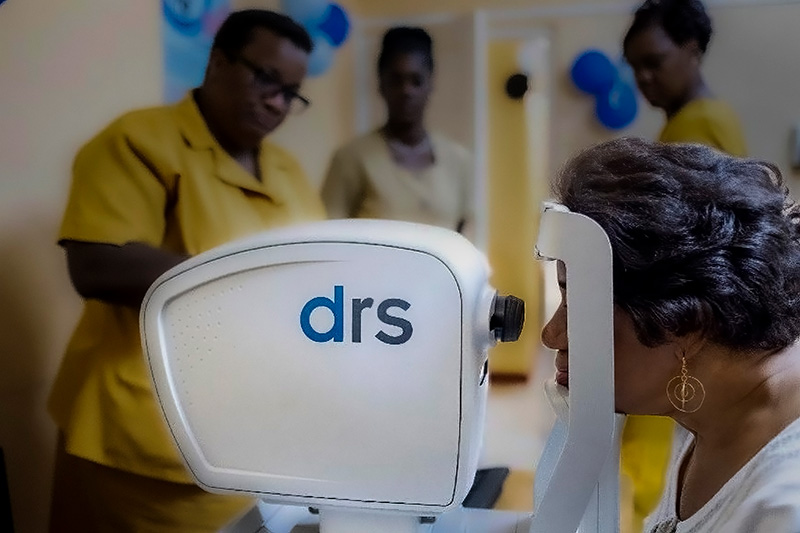 A female patient sits with her chin resting on the chin rest of the camera. Tehre are 3 eye care staff members standign in the background of the photo.