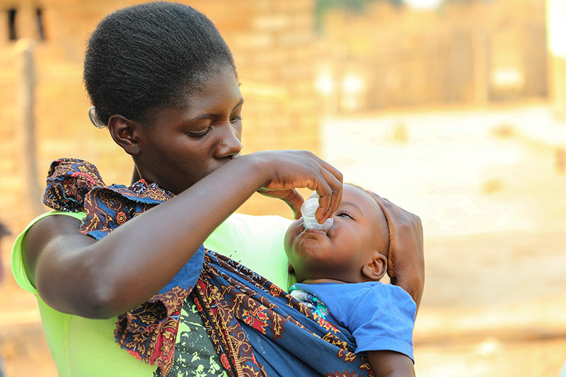 A mother gives her a young child a does of medicine with a small white cup, pouring it into the child'smouth