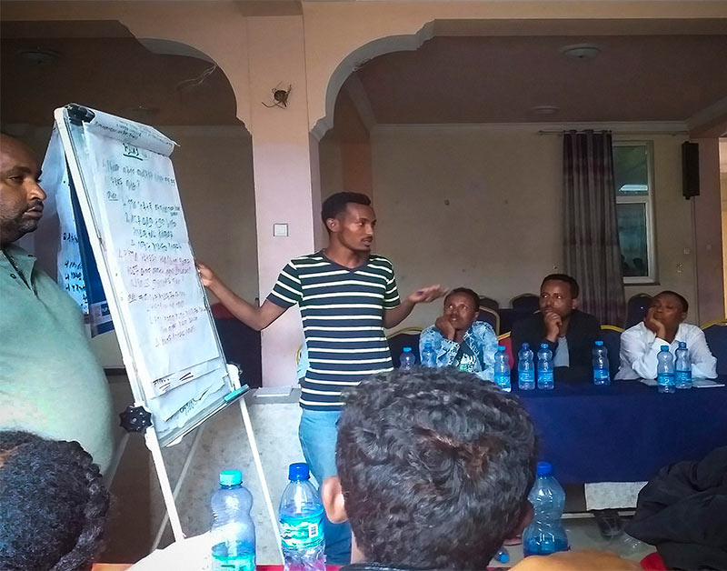 A man stands next to a flip chart among other people in a room, to whom he is talking.