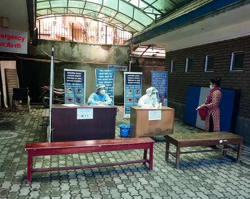 Two eye care workers sitting behind desks in a hospital area, both wearing full PPE and a female patient wearing a mask, approaching the desk