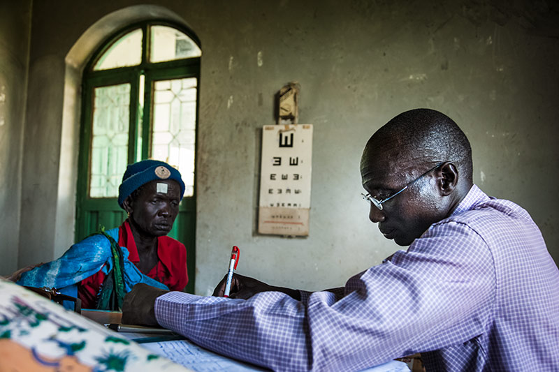 A male ophthalmic staff member sitting at a desk writing with a female patient sitting opposite him and a tumbling E chart hanging on the wall in the background.