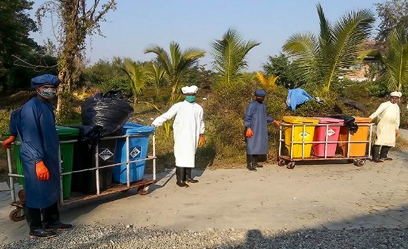 Four hospital staff members wearing PPE pull trolleys loaded with waste in different colour coded bins