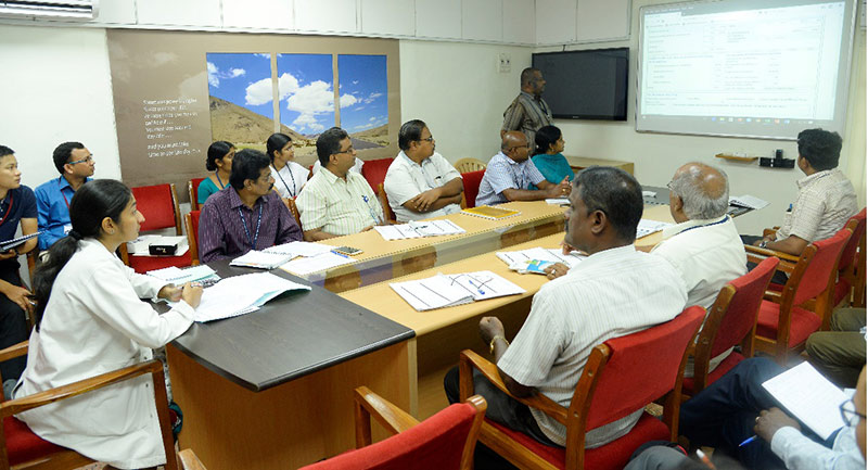 Hospital staff sitting round a large table listening to a presentation, looking. at a screen