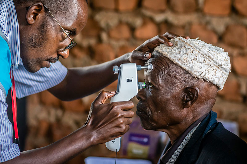 An eye care worker holds a portable screening device against the face of an African gentleman with his right hand, while using his left hand to steady the head of the patient.
